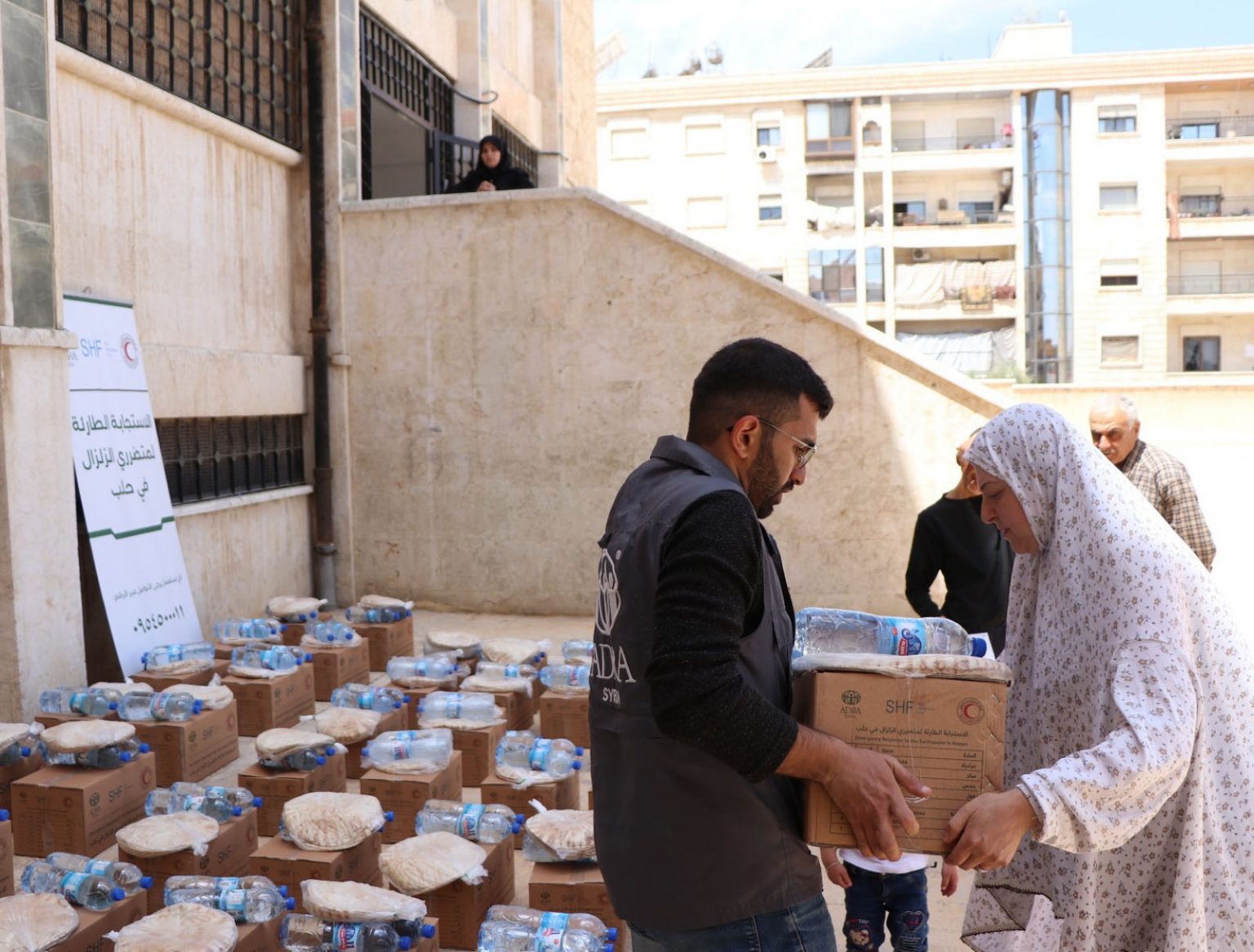 ADRA-Helfer überreicht syrischer Frau Hilfspaket mit Wasser und Fladenbrot – umgeben von weiteren gepackten Kartons