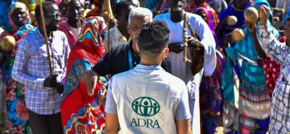 ADRA-Mitarbeiter steht vor bunt gekleideter Menschenmenge in Afrika – Versammlung mit Musik und Gesprächen im Freien