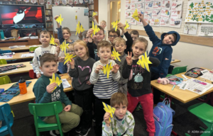 Schulklasse hält gelbe Papierfische hoch, viele Kinder lächeln.