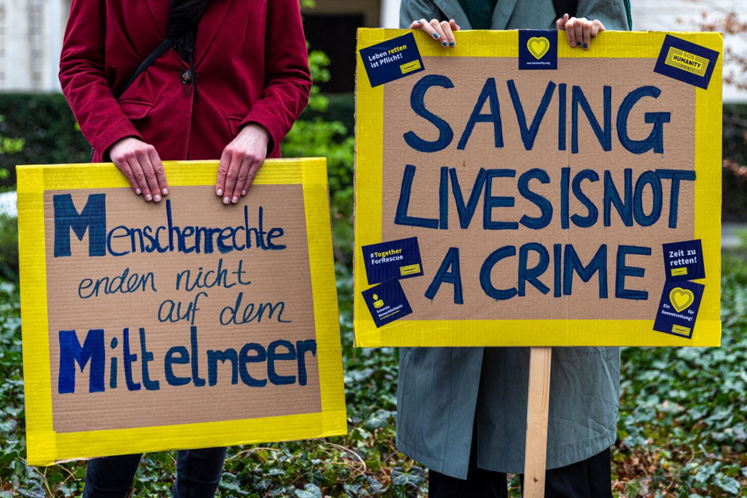 Zwei Personen mit Protestschildern „Menschenrechte enden nicht auf dem Mittelmeer“ und „Saving lives is not a crime“. © SOS Humanity/Wanda Proft