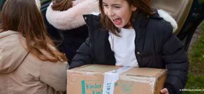 Mädchen trägt ein Hilfspaket mit dem Logo der Aktion „Kinder helfen Kindern!“ und lächelt freudig.