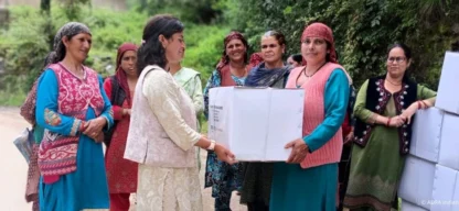 Humanitäre Hilfe in Himachal Pradesh: Verteilung von Hilfsgütern: Zwei Frauen halten einen großen weißen Karton, neben ihnen warten weitere Frauen; Kartons sind am Wegesrand gestapelt