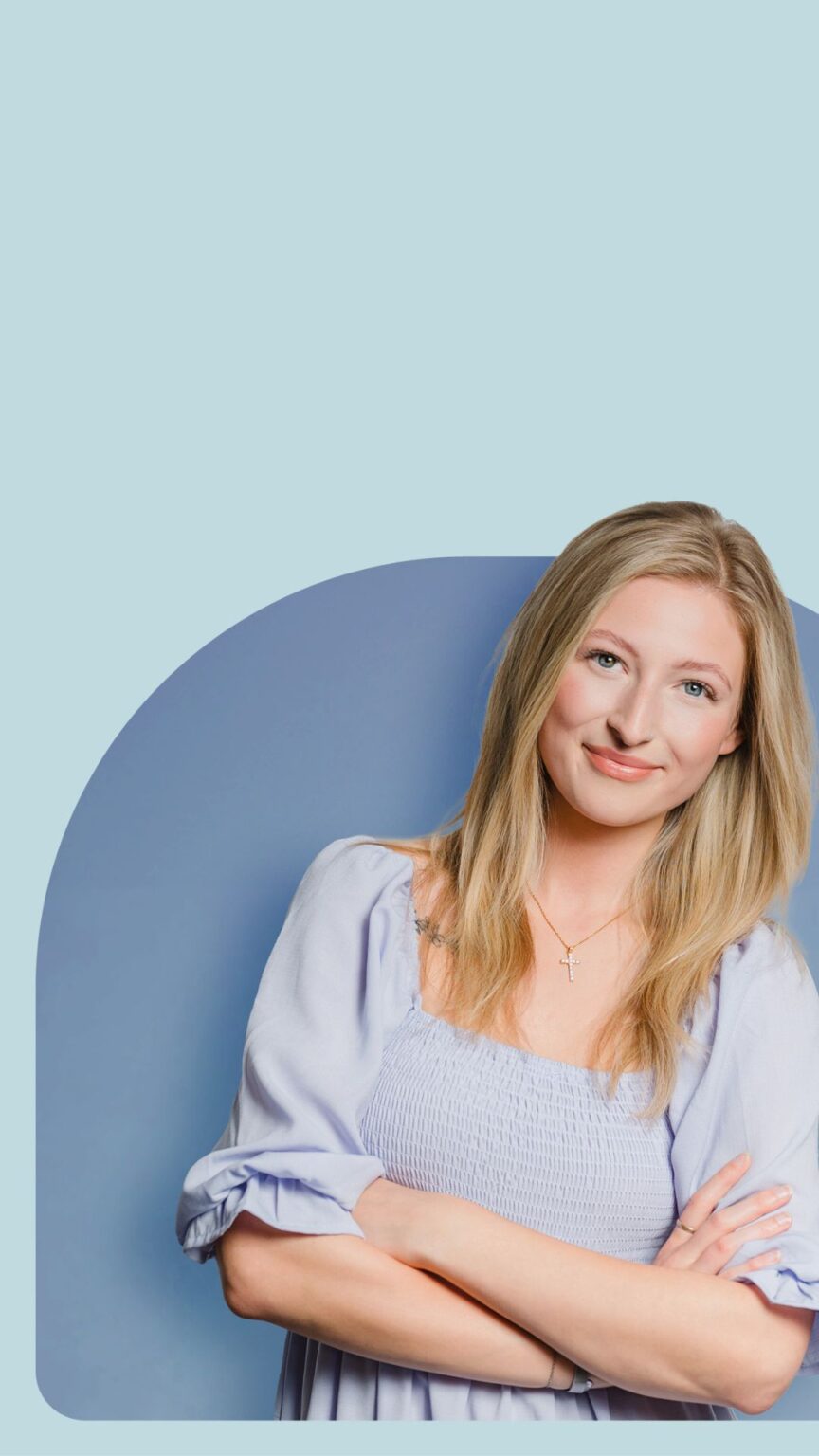 Frau mit langen blonden Haaren und hellblauer Bluse steht vor blauem Hintergrund und lächelt mit verschränkten Armen.
