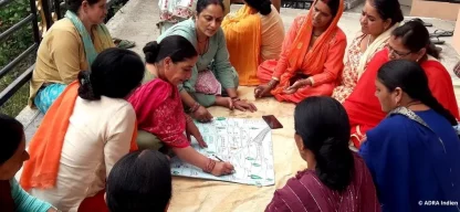 Eine Gruppe von Frauen aus Indien sitzt im Kreis auf dem Boden und diskutiert über ein großes Blatt Papier mit Diagrammen. Sie scheinen gemeinsam an einer Planung oder Schulung teilzunehmen.