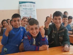 Gruppe von Kindern in einem Klassenraum, drei Kinder sitzen vorne an einem Holztisch und zeigen mit lächelnden Gesichtern den Daumen nach oben.