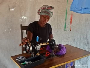 Eine Frau sitzt an einem Tisch und näht Stoff an einer Nähmaschine in einem Raum.