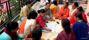 Eine Gruppe von Frauen sitzt im Kreis, diskutiert und arbeitet gemeinsam an einem großen Papierbogen mit Zeichnungen und Plänen auf einer Terrasse in Indien