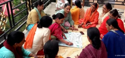 Eine Gruppe von Frauen sitzt im Kreis, diskutiert und arbeitet gemeinsam an einem großen Papierbogen mit Zeichnungen und Plänen auf einer Terrasse in Indien