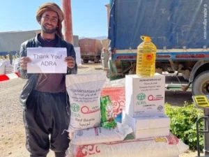 Ein Mann steht im Freien vor Hilfsgütern wie Mehl, Öl und Bohnen und hält ein Schild mit einem Dankeschön-Text hoch.