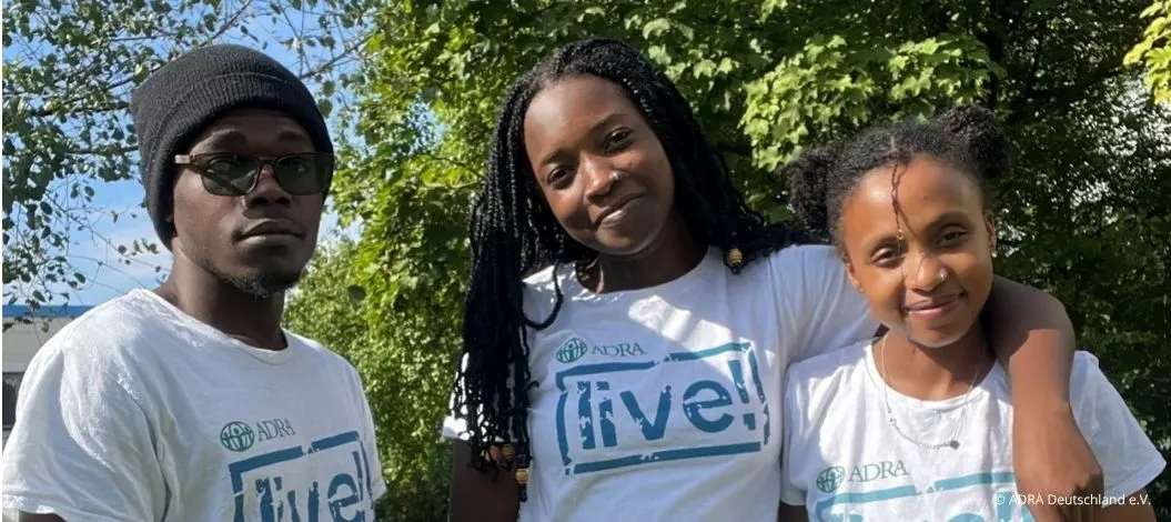 Drei junge Personen stehen draußen im Grünen vor Bäumen. Alle tragen weiße ADRAlive! T-Shirts. Die Person links trägt eine Mütze und Brille, die mittlere Person lächelt und hat lange geflochtene Haare, und die Person rechts lehnt sich an die mittlere und lächelt ebenfalls.