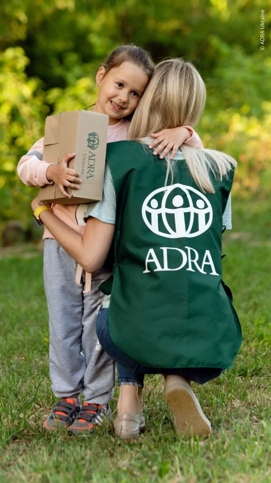 Ein Kind umarmt eine ADRA Mitarbeiterein in einer grünen ADRA-Weste und hält dabei ein Hilfspaket, beide stehen auf einer Wiese.