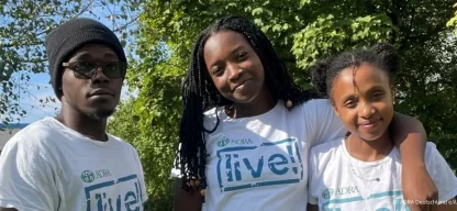 Drei junge Personen stehen draußen im Grünen vor Bäumen. Alle tragen weiße ADRAlive! T-Shirts. Die Person links trägt eine Mütze und Brille, die mittlere Person lächelt und hat lange geflochtene Haare, und die Person rechts lehnt sich an die mittlere und lächelt ebenfalls.