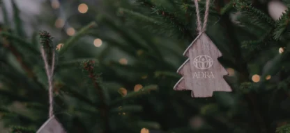 Holzanhänger in Form eines kleinen Weihnachtsbaums mit ADRA-Logo hängt an einem Tannenzweig; unscharfe Lichter im Hintergrund sorgen für eine warme, festliche Stimmung.