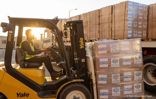 Ein Mitarbeiter lädt mit einem Gabelstapler palettierte Hilfspakete mit ADRA- und Plan-International-Logos auf einen LKW.
