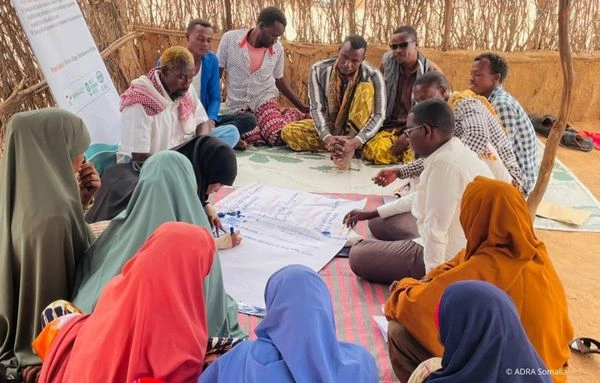 Eine Gruppe von Menschen sitzt in einem Kreis in einer einfachen, offenen Hütte und arbeitet gemeinsam an einem großen Papierbogen, um lokale Maßnahmen zur Katastrophenvorsorge zu planen.