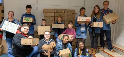 Kinder, Jugendliche und Erwachsene stehen gemeinsam in einem Raum und halten Geschenkpakete der Aktion ›Kinder helfen Kindern‹ in den Händen. Im Hintergrund sind viele weitere Pakete gestapelt. Die Gruppe lächelt in die Kamera