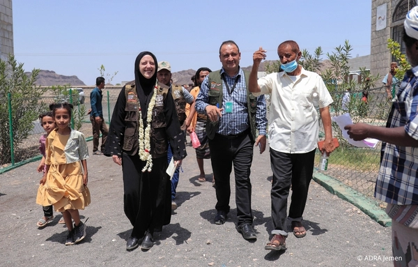 Eine Gruppe von Menschen geht gemeinsam durch ein Gelände im Jemen, darunter eine Verena Lauer in ADRA-Kleidung und lokale Begleitpersonen.