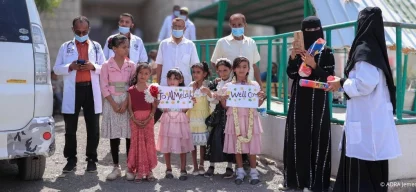 Mehrere Kinder stehen vor einer Gesundheitseinrichtung im Jemen und halten selbstgemalte Willkommensschilder. Hinter ihnen stehen medizinische Mitarbeitende, einige tragen Schutzmasken.