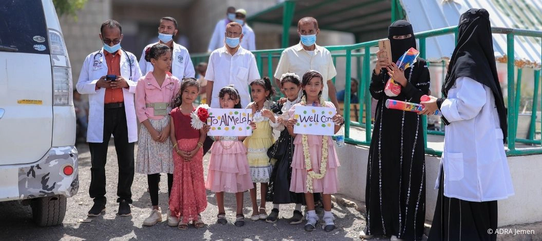Mehrere Kinder stehen vor einer Gesundheitseinrichtung im Jemen und halten selbstgemalte Willkommensschilder. Hinter ihnen stehen medizinische Mitarbeitende, einige tragen Schutzmasken.