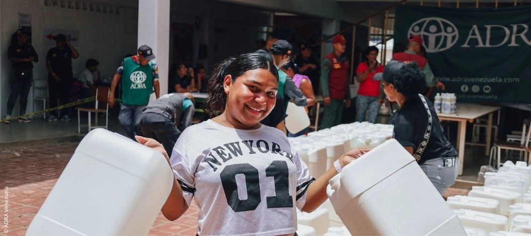 Eine lächelnde Frau aus Venezuela trägt zwei leere Wasserkanister, während ADRA-Helferinnen und -Helfer im Hintergrund Hilfsgüter verteilen