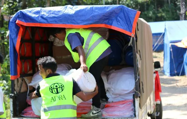 Zwei ADRA-Helfer in Warnwesten laden Säcke mit Hilfsgütern aus einem Fahrzeug in einem Zeltlager in Nepal aus