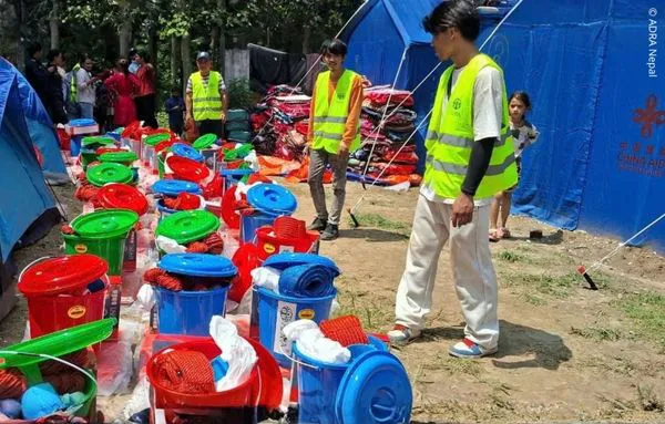 ADRA-Helfer stehen zwischen Zelten und bereitgestellten Hilfsgütern wie Eimern, Decken und weiteren Haushaltsartikeln