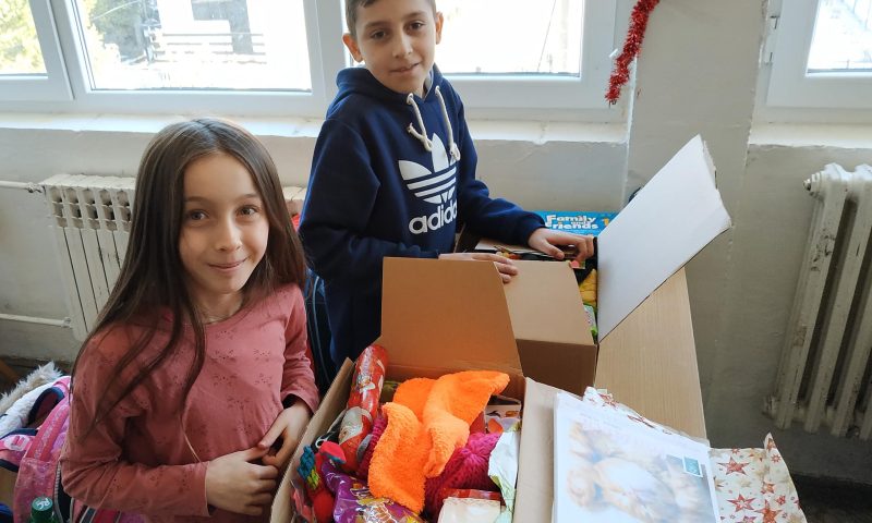 Ein junge und ein Mädchen packen gemeinsam KHK Pakete aus, welche aus Süßigkeiten, Spielzeuge, Kuscheltiere und Klamotten bestehen