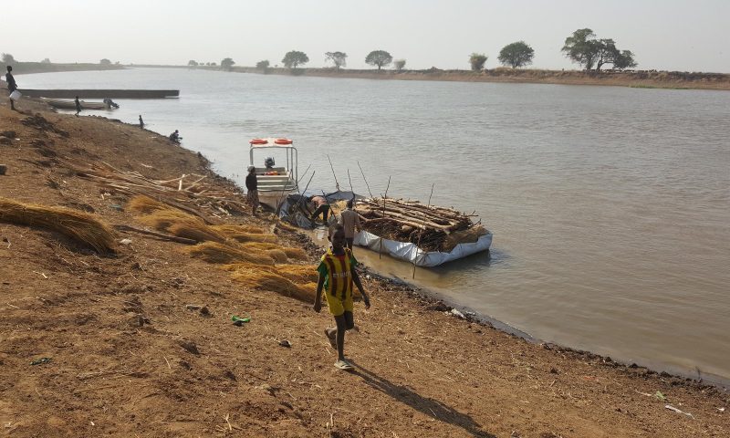 Ein Junge läuft am Ufer eines Flusses, dahinter ist ein Floss, mit dem Baumstämme und Holz transportiert werden