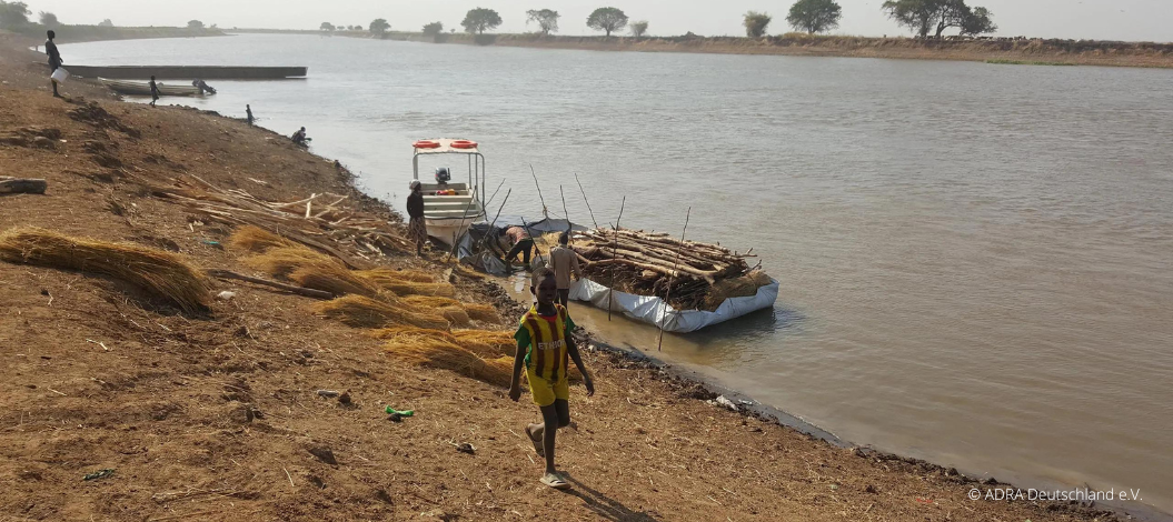 Ein Junge läuft am Ufer eines Flusses, dahinter ist ein Floss, mit dem Baumstämme und Holz transportiert werden