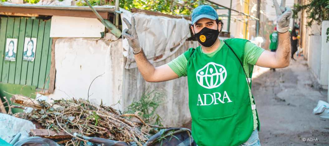 ADRA-Mitarbeiter in grüner Weste, blauer Basecap, Handschuhen und Smiley-Mundschutz hebt die Arme und zeigt das Peace-Zeichen