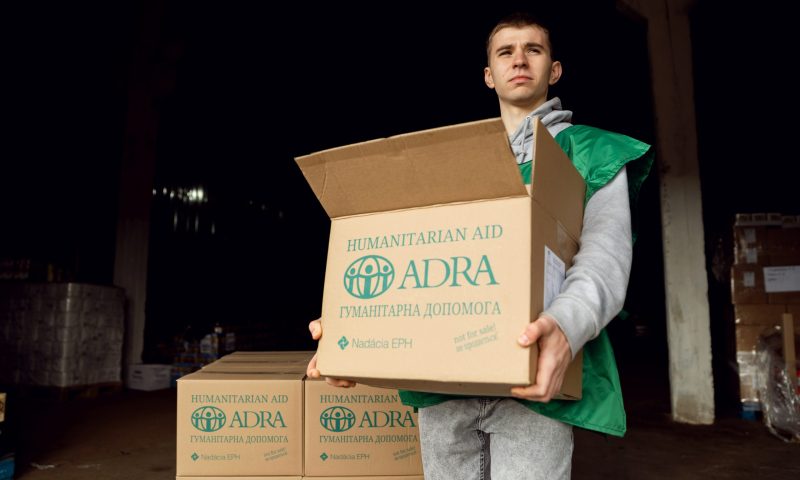 Ein ADRA-Mitarbeiter steht in einem Lager und hält ein Paket für humanitäre Unterstützung mit beiden Händen. Im Hintergrund stehen weitere Hilfspakete.