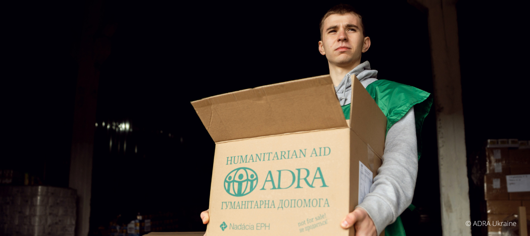 Ein ADRA-Mitarbeiter steht in einem Lager und hält ein Paket für humanitäre Unterstützung mit beiden Händen. Im Hintergrund stehen weitere Hilfspakete.