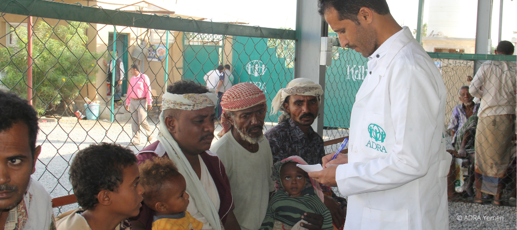 ADRA-Mitarbeiter im weißen Kittel spricht mit wartenden Männern und Kindern in einer Einrichtung im Jemen