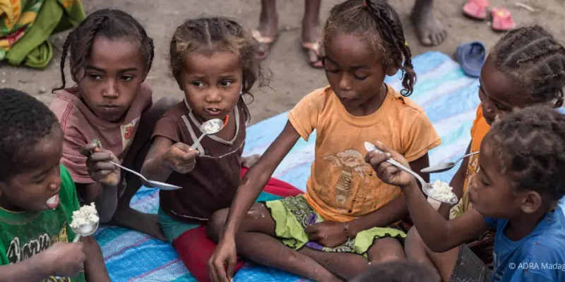 Eine Gruppe hungernde Kinder und Kleinkinder sitzt um eine Schüssel mit Brei gefüllt und teilt sich das Essen
