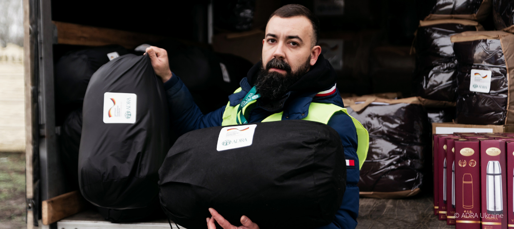 ChatGPT: Ein ADRA-Mitarbeiter in gelber Warnweste hält zwei schwarze Hilfspakete mit ADRA-Logo vor einem Transporter voller weiterer Hilfsgüter