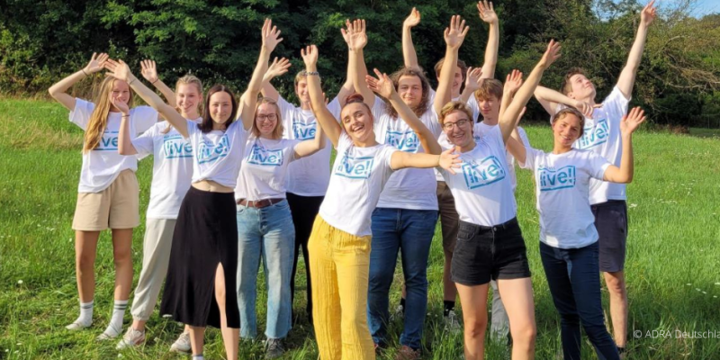 Eine lebendige Szene zeigt eine Gruppe von ADRAlive!-Freiwilligen in strahlend weißen T-Shirts, die sich auf einer üppig grünen Wiese versammelt haben. Mit einem begeisternden Lächeln auf ihren Gesichtern heben sie gemeinsam ihre Arme in die Luft, während das ADRAlive!-Logo stolz auf ihren Brustbereichen prangt