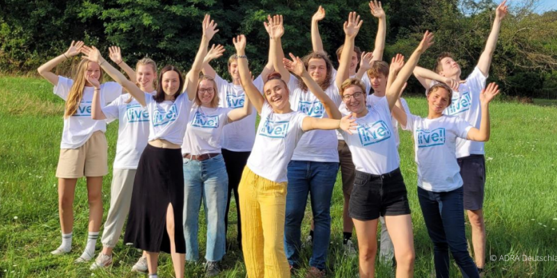ADRAlive!-Freiwillige in weißen T-Shirts heben lächelnd auf grüner Wiese die Arme