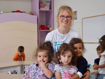 Eine lächelnde Mitarbeiterin von ADRAlive hält drei kleine Mädchen vor sich. Im Hintergrund befinden sich drei weitere Kinder