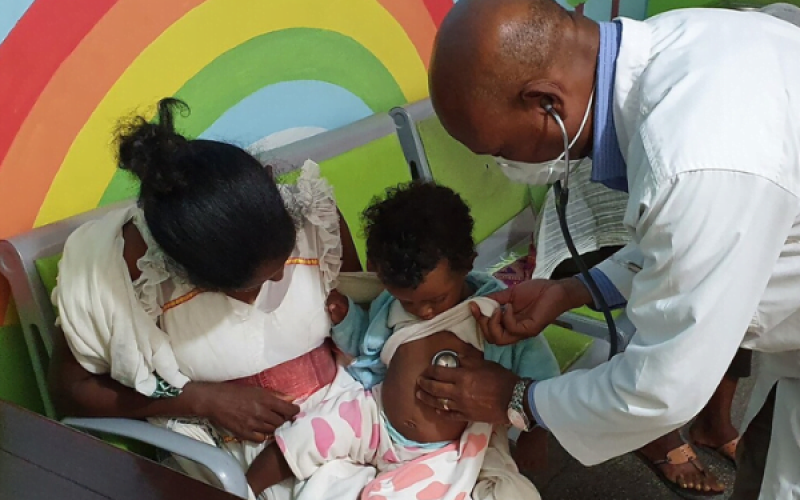 Äthiopien_PRO2720108_Kinderklinik Ein Kleinkind sitzt auf dem Schoß der Mutter vor einer Regenbogenwand, während ein Arzt im weißen Kittel, das Kind mit einem Stethoskop untersucht