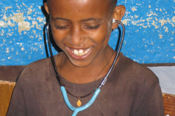 Ein lächelnder Junge sitzt auf einem Stuhl und untersucht sich mit einem Stethoskop, welches er in beiden Ohren trägt