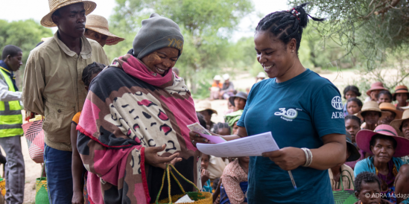 ADRA-Mitarbeiterin übergibt einer Frau in Madagaskar Unterlagen, im Hintergrund warten Menschen auf Hilfsgüter