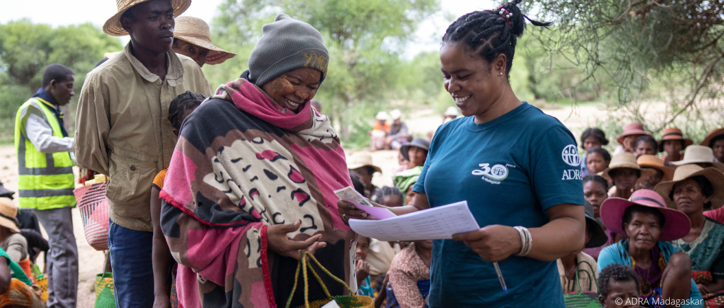 ADRA-Mitarbeiterin übergibt einer Frau in Madagaskar Unterlagen, im Hintergrund warten Menschen auf Hilfsgüter