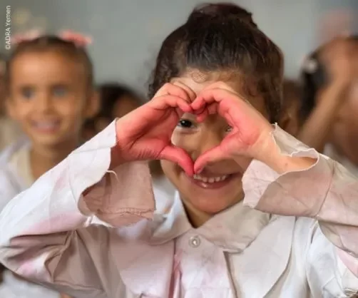 Mädchen in jemenitischer Schulklasse formt mit rot bemalten Händen ein Herz vor dem Gesicht und blickt hindurch
