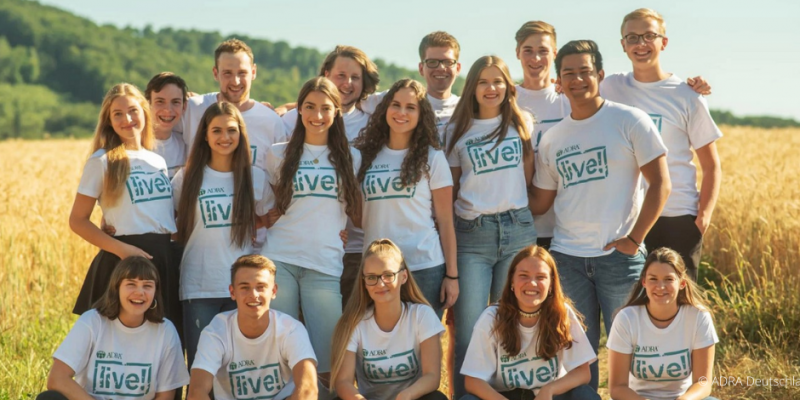 Gruppenfoto von ADRAlive!-Freiwilligen auf einem Feld: Eine Gruppe von Freiwilligen steht lächelnd auf einem offenen Feld, umgeben von grüner Landschaft. Sie tragen ein weißes T-Shirt mit dem Logo von ADRAlive! und sind in guter Stimmung.
