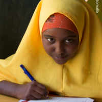 Mädchen in gelbem Kopftuch schreibt in ein Heft und schaut in die Kamera
