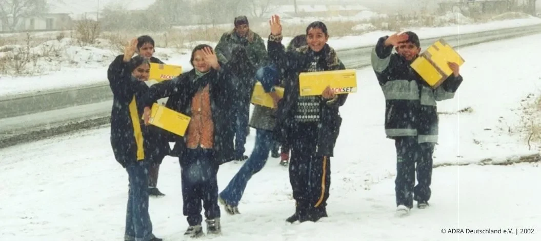 Kinder in Bulgarien winken im Schneefall und tragen gelbe Hilfspakete (ADRA Deutschland e.V., 2002).