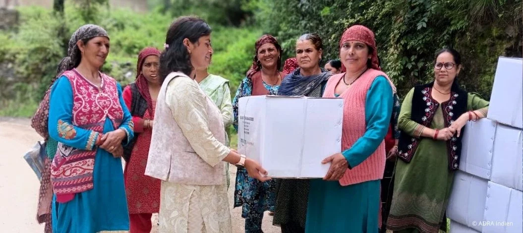 Humanitäre Hilfe in Himachal Pradesh: Verteilung von Hilfsgütern: Zwei Frauen halten einen großen weißen Karton, neben ihnen warten weitere Frauen; Kartons sind am Wegesrand gestapelt