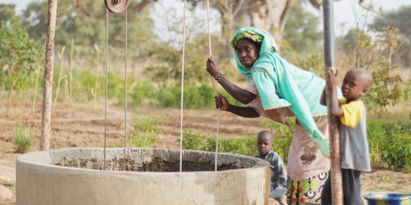 Frau aus Mali zieht Wasser aus Brunnen, begleitet von zwei kleinen Kindern – alle blicken in die Kamera