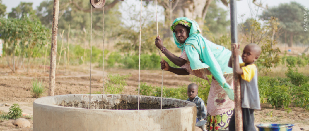 Frau aus Mali zieht Wasser aus Brunnen, begleitet von zwei kleinen Kindern – alle blicken in die Kamera