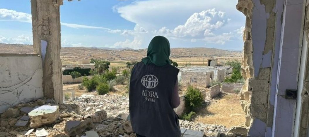 ADRA-Mitarbeiterin steht im Freien vor zerstörten Gebäuden in Syrien nach dem Erdbeben 2023. | ©ADRA Syrien